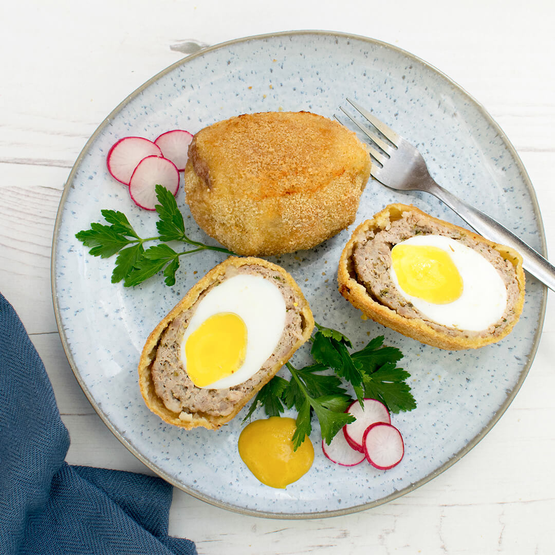 A plate with a whole and halved scotch egg, served with fresh herbs, radish and mustard