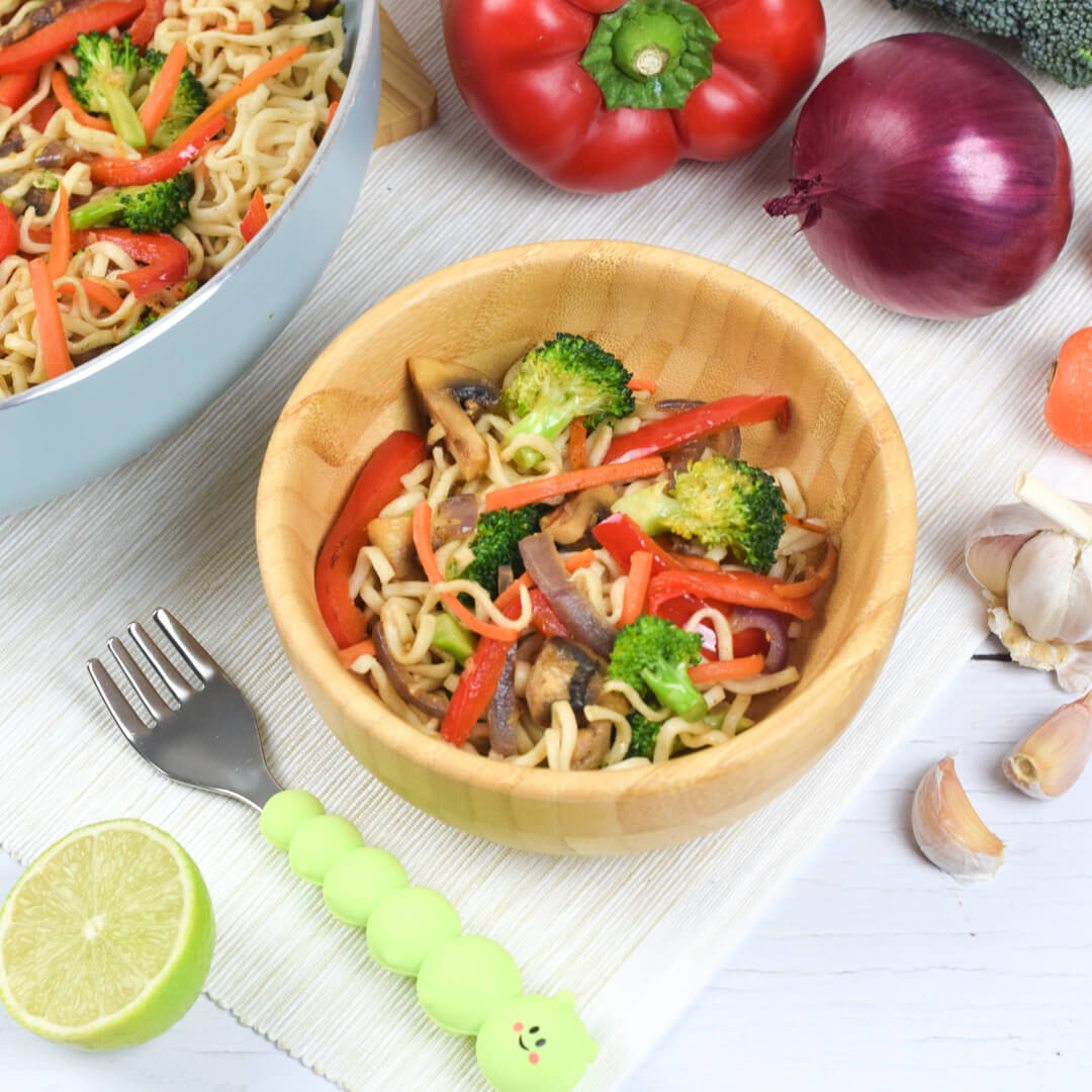 A baby bamboo bowl with vegetable stir fry inside. Larger Bowl with Stir fry next to it. Also vegetables around the bowls. 