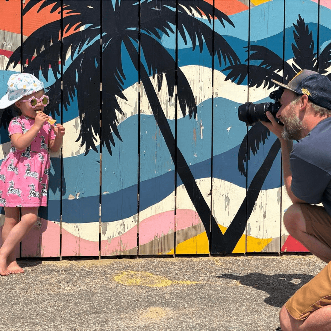 Toddler dressed in pink dress wearing a sun hat and sun glasses and eating an Organix oaty bar while leaning on a summery wall with a the image of a beach. An adult is taking a picture of the toddler. 