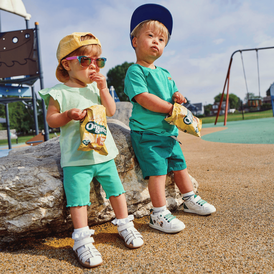 Two children at a play area dressed in bright green clothes, wearing colourful sun hats and sun glasses in hip hop style and eating Organix snacks