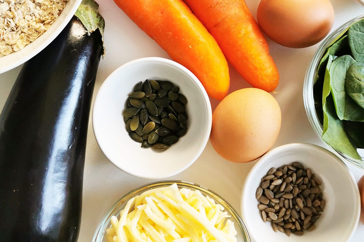 A bowl of oats next to a bowl of spinach, pumpkin seeds, sunflower seeds, grated cheese, 2 eggs, carrots and an aubergine