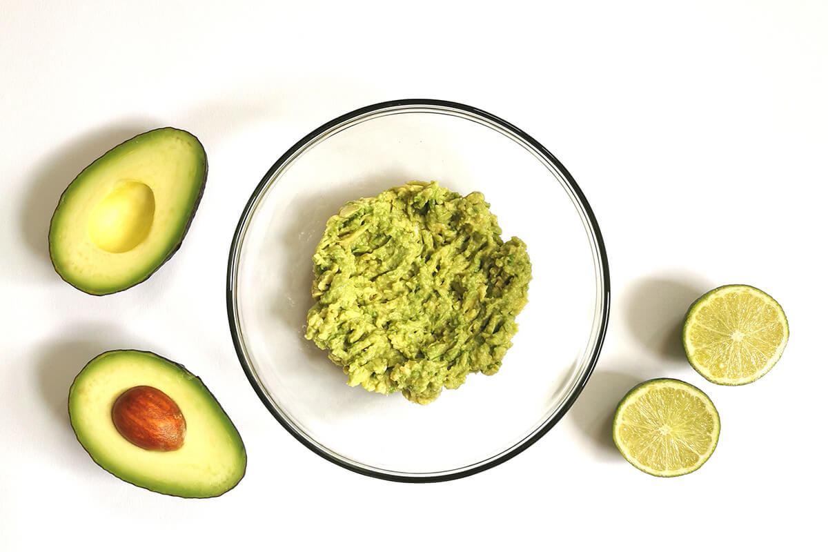 A glass bowl with smashed avocado next to a halved avocado and halved lime