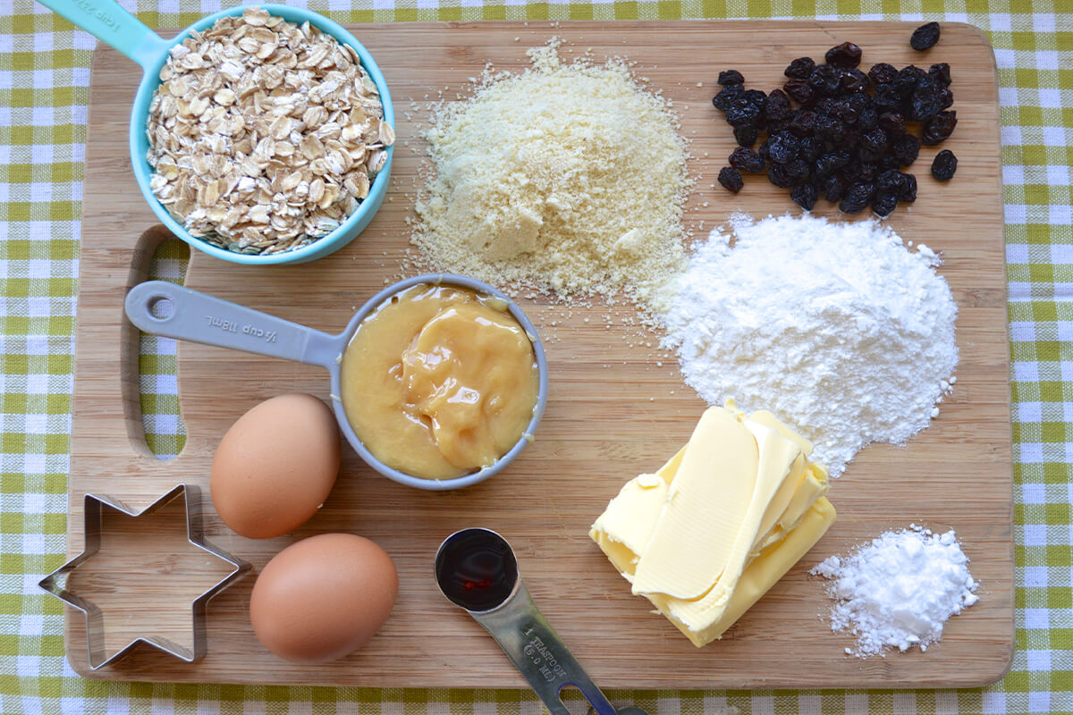 A chopping board with oats, ground almonds, butter, rice flour, eggs, raisins, honey, baking powder, vanilla extract and a star shaped cookie cutter