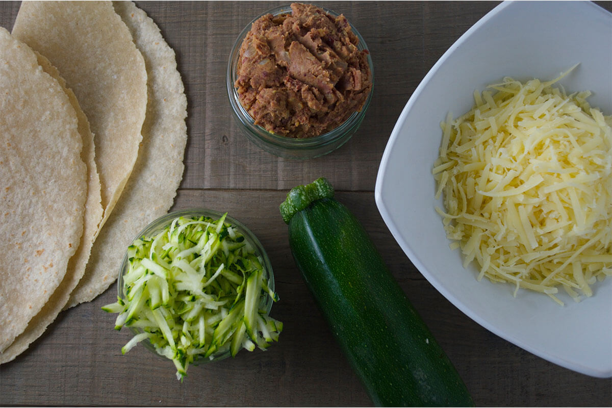 A table with some wraps, red kidney bean spread in a ramekin, grated courgette in a ramekin next to a whole courgette and some grated cheese in a white bowl