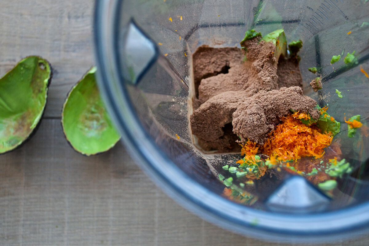 A food processor with avocado, orange zest, cocoa powder, banana and raw coconut oil