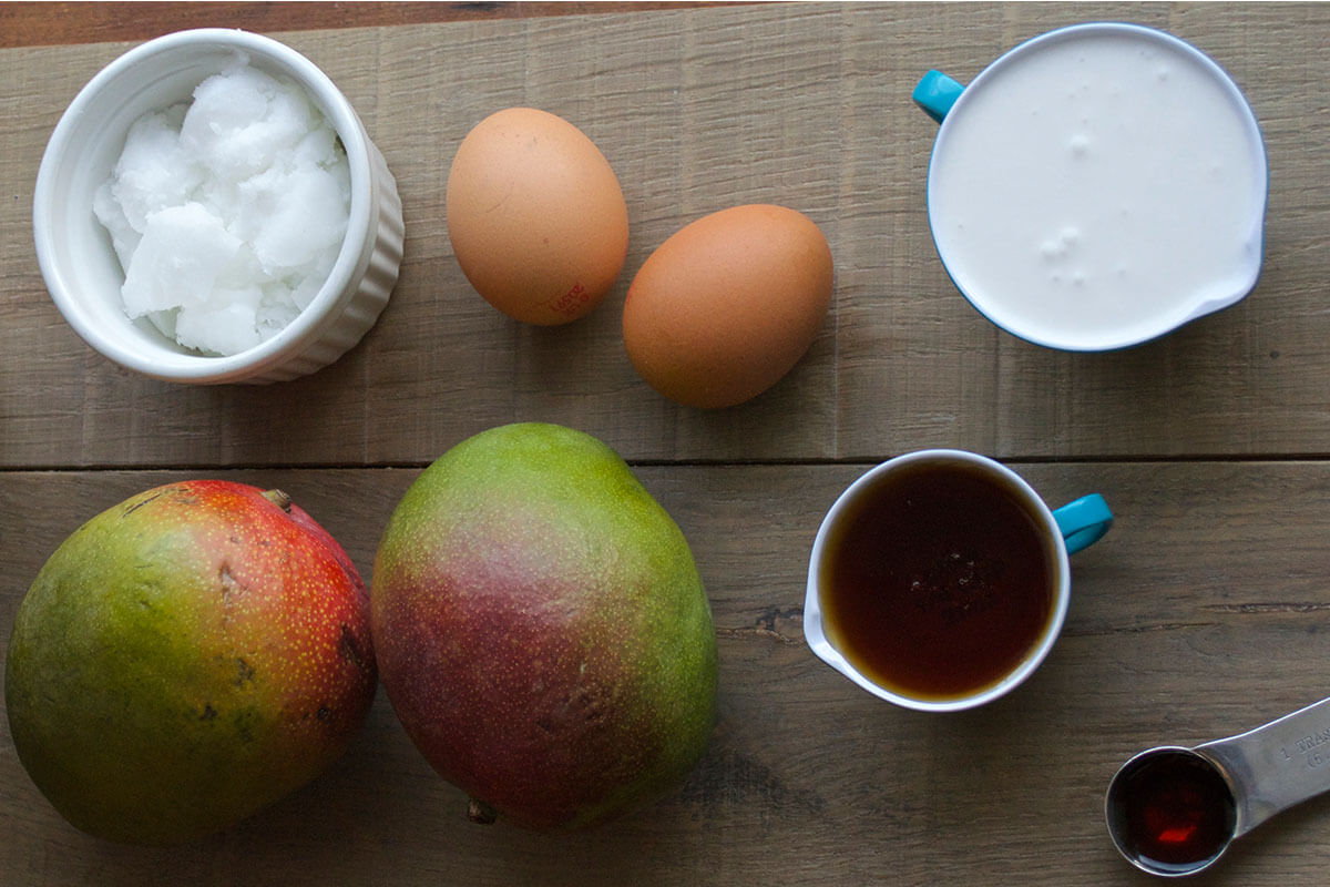 A table with 2 mangos, 2 eggs, a ramekin of coconut oil, some maple syrup and coconut cream