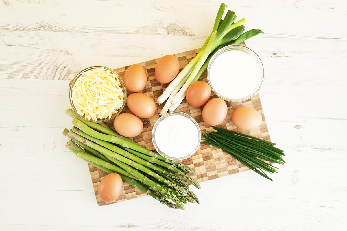 Asparagus, spring onions, eggs, water and milk, chives, grated cheese and soft goats cheese on a chopping board