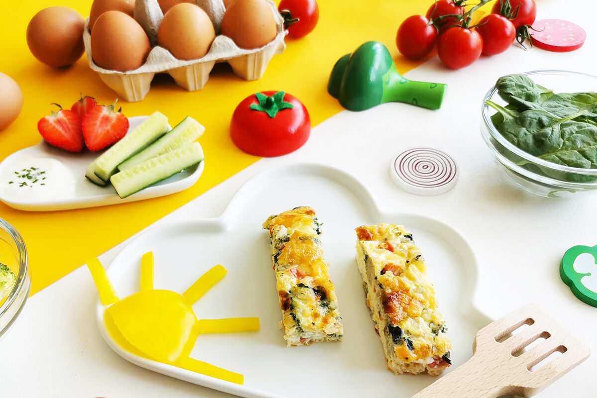 A serving of omelette fingers next to a case of 6 eggs, some spinach leaves, tomatoes, cucumber sticks and strawberries