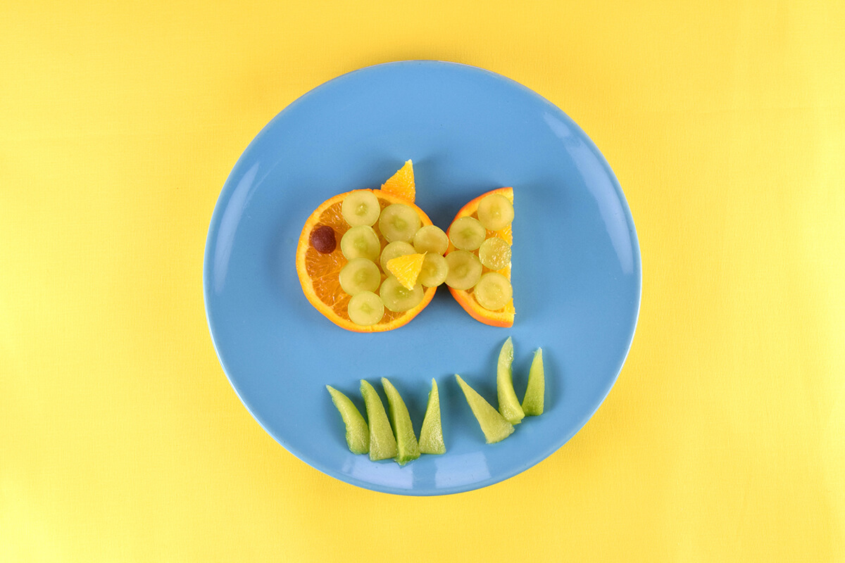 Sliced grapes placed on top of oranges to create scales. Strawberry and apple gummie placed at front orange to create an eye. Triangles of orange placed on grapes to create fins