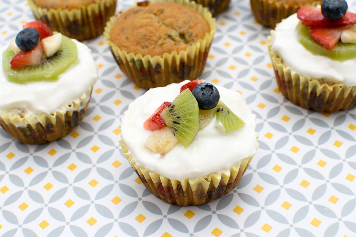 A plate of Gummy banana cupcakes, some un-topped/undecorated and some decorated with Greek yoghurt and fruit segments/slices