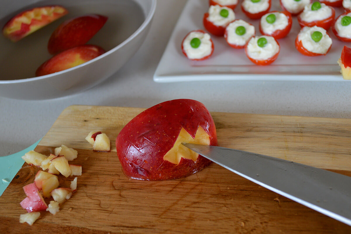 A chopping board with apple cut to look like a mouth with sharp teeth, next to a platter with cream cheese stuffed cherry tomatoes topped with peas