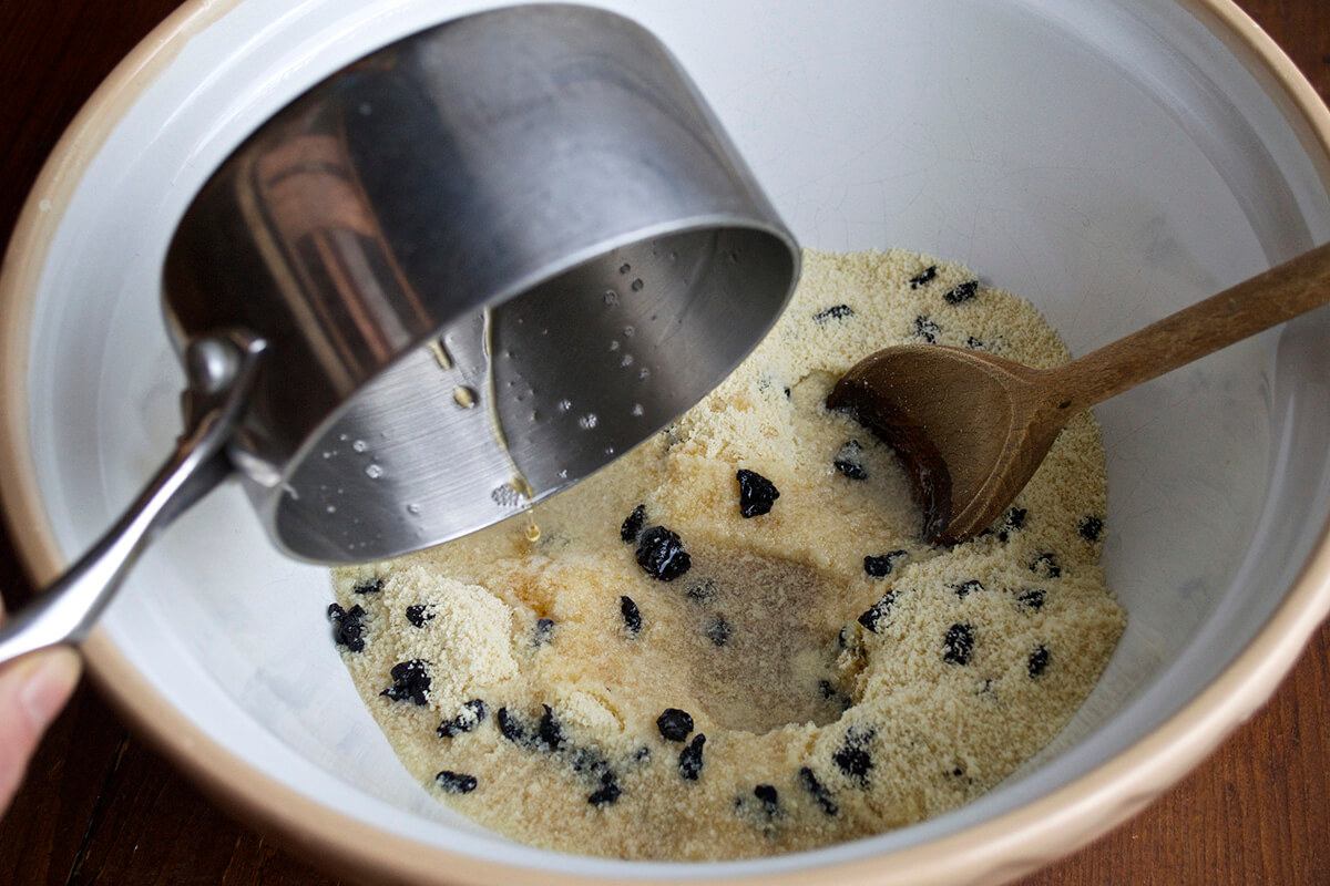 A large bowl of ground almonds and sour cherries with maple syrup and vanilla extract being added