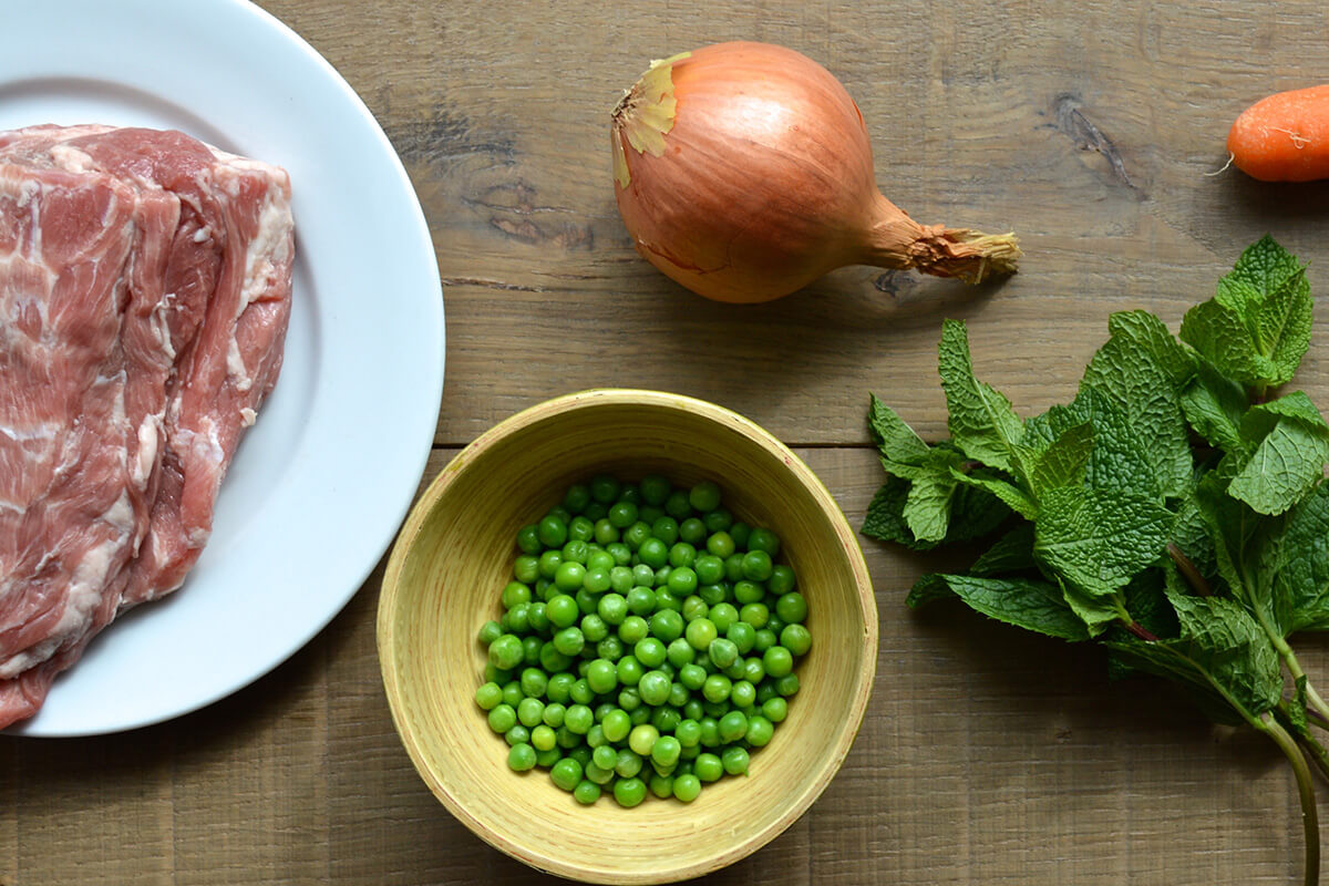 Lamb fillet on a plate next to a bowl of peas, an onion, carrot and some mint leaves