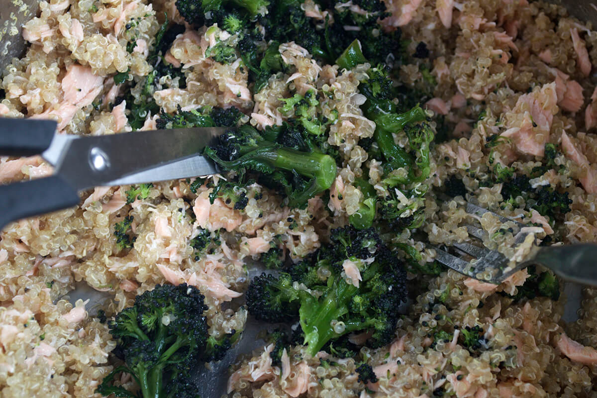 Quinoa, salmon and purple sprouting broccoli in a pan while the broccoli is being cut with scissors