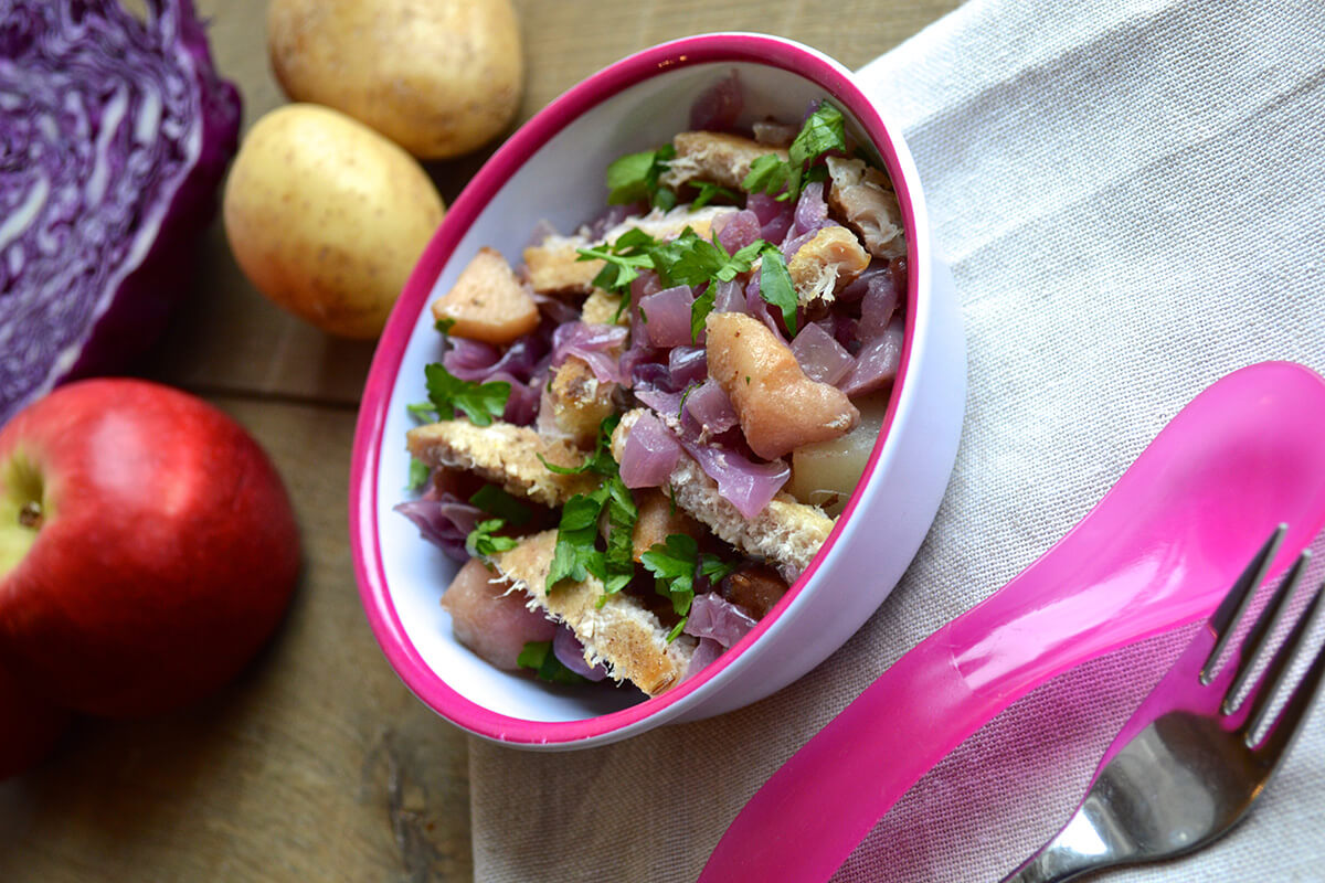 A small bowl of Slow Cooked Red Cabbage & Pork next to an apple, 2 potatoes and a halved red cabbage